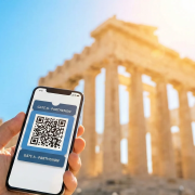 A high-resolution, photorealistic travel image taken from a low angle looking up. In the foreground, a traveler's hand is holding a modern smartphone displaying a digital ticket with a QR code clearly visible on the screen. The focus is on the phone and the hand. In the background, slightly soft-focused (bokeh effect), stands the majestic Parthenon under a brilliant clear blue Greek sky. The sun is shining, creating a "lens flare" effect that suggests a bright, happy morning. The image should convey success, ease of entry, and modern travel convenience. Style: Professional travel photography, 8k resolution, vibrant colors.